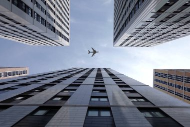 Big airplane flying high above modern city skyscraper buildings with many windows in business cluster view bottom up on bright sunny day clipart