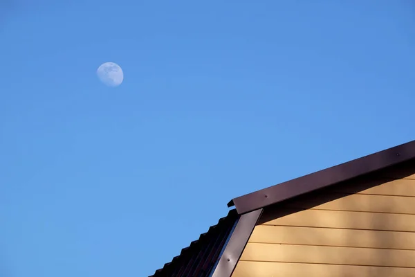 晴れた日のフロントビューのクローズアップでは 澄んだ青い雲のない空に黄色の側面と茶色の金属屋根と丸い月で覆われた田舎の家の壁の一部 — ストック写真