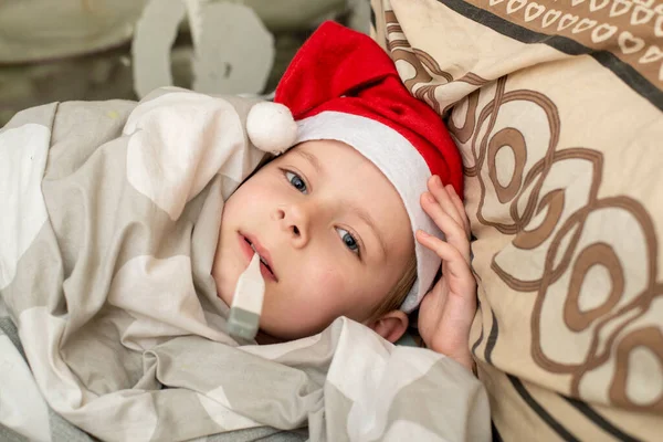サンタの帽子の少年は病気です ベッドの温度を測定します 新年の病気 — ストック写真