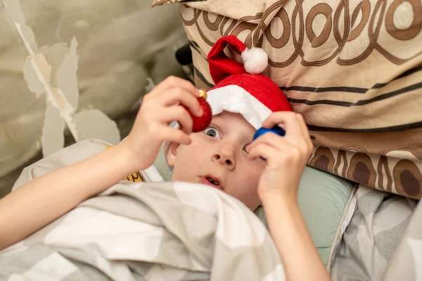 クリスマスだ サンタの帽子の男の子はクリスマスの装飾とふけるベッドに横たわっています — ストック写真