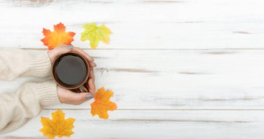 Sıcak süveter giymiş kadınların elleri bir fincan tutuyor. Beyaz ahşap arka planda sonbahar yaprakları. Pankart.