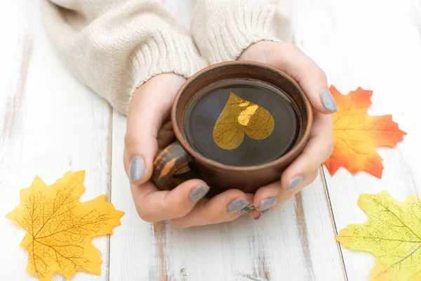 Women\'s hands in a warm sweater are holding a cup with heart. Autumn leaves on a white wooden background.