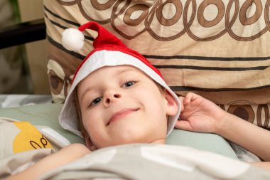 Tatmin olmuş, gülümseyen bir çocuk Noel Baba şapkasıyla yatakta yatıyor. Tatillerin neşeli hissi - Yeni yıl, Noel.