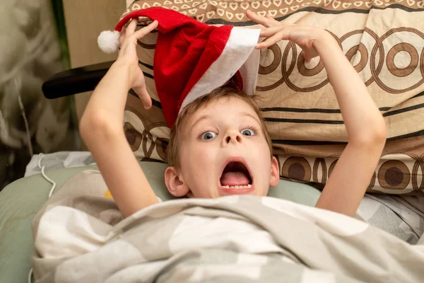 クリスマスだ サンタの帽子の男の子がベッドに横になって顔を作っている — ストック写真