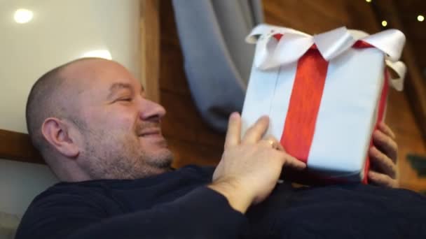 Άνθρωπος Χαμογελάει Χαίρεται Και Κρατά Ένα Δώρο Ξαπλωμένος Στον Καναπέ — Αρχείο Βίντεο