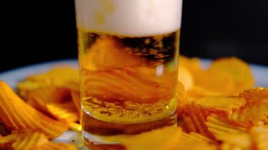 Bira koyu bir arka planda bir bardağa dökülür. Cips etrafa saçılmış..