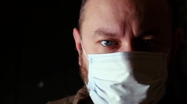 Tıbbi maskeli bir adam siyah arka plandaki kameraya yakından bakar..