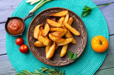 Tabakta büyük parçalar halinde patates kızartması. Masada turkuaz peçete ve farklı sebzeler var..