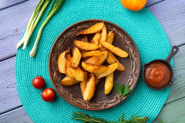 Tabakta büyük parçalar halinde patates kızartması. Masada turkuaz peçete ve farklı sebzeler var..