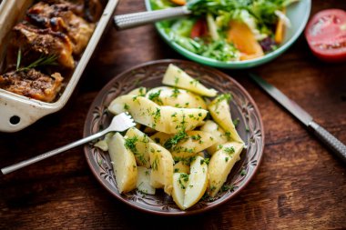 Dereotlu haşlanmış patates, taze sebze salatası ve koyu ahşap arka planda fırında tavuk butları..