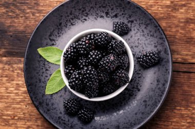 Taze böğürtlenler, beyaz bir kasede, siyah bir tabakta, ahşap bir arka planda..