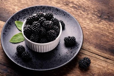 Taze böğürtlenler, beyaz bir kasede, siyah bir tabakta, ahşap bir arka planda..