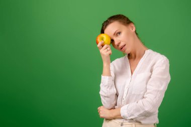 Beyaz gömlekli orta yaşlı güzel bir kadın yeşil arka planda bir elma tutuyor. Sağlıklı beslenme, vejetaryenlik, sağlıklı görünüm.