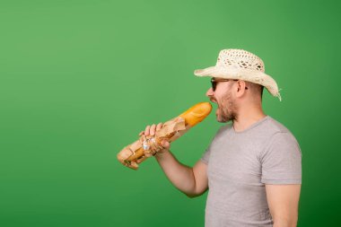 Şapkalı ve güneş gözlüklü bir adam yeşil bir arka planda Fransız ekmeğini ısırıyor..