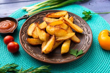 Tabakta büyük parçalar halinde patates kızartması. Masada turkuaz peçete ve farklı sebzeler var..