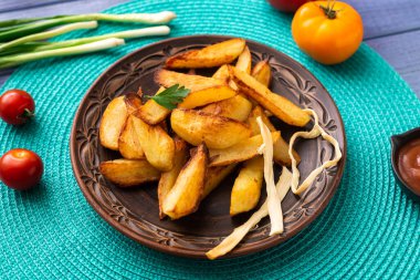 Tabakta büyük parçalar halinde patates kızartması. Masada turkuaz peçete ve farklı sebzeler var..