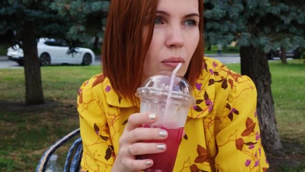 一位穿着黄色衬衫的红头发女人坐在长椅上休息着 喝着街上塑料杯子里的鸡尾酒 — 图库视频影像