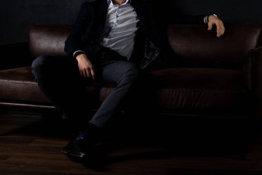 Tarz sahibi kıyafetleri içinde tanınmayan bir adam karanlıkta kanepede rahatça oturur..