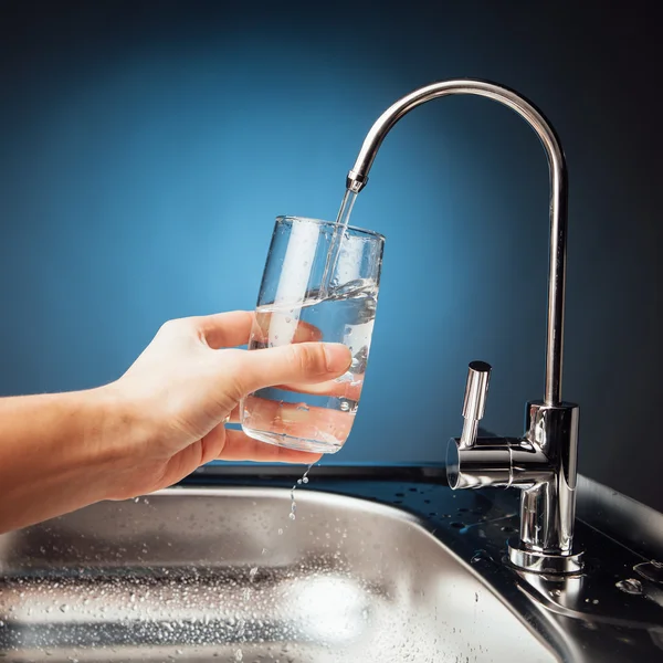 Mano versando un bicchiere d'acqua dal rubinetto del filtro, fondo blu — Foto Stock