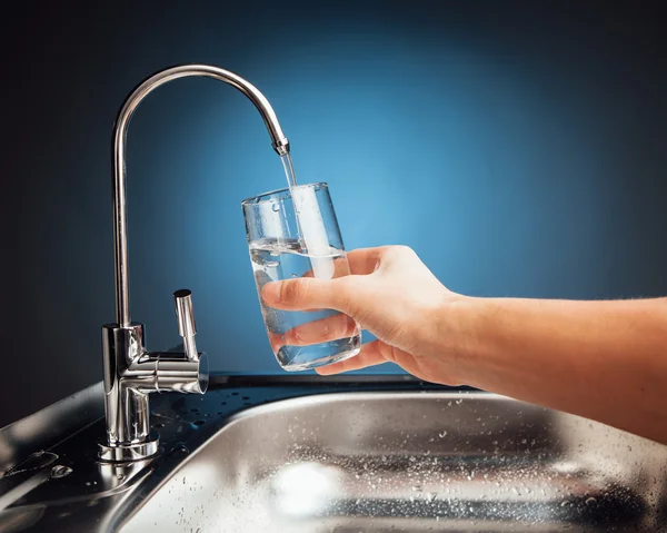 Mano versando un bicchiere d'acqua dal rubinetto del filtro, fondo blu — Foto Stock