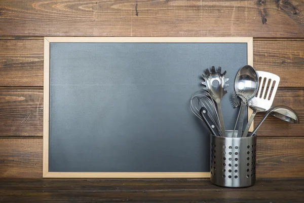 Wooden background with a chalkboard and cooking utensils — Stock Photo, Image