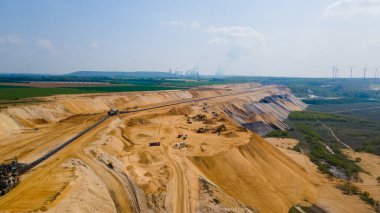 Büyük bir linyit madeninin yüksek açılı insansız hava aracı görüntüsü. Ağır madencilik makineleri, yeşil alanlar, rüzgar türbinleri ve açık bir gökyüzü altında termik santrallerin yer aldığı sanayi sahnesi