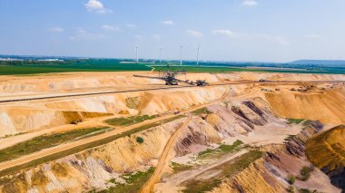 Büyük bir linyit madeninin yüksek açılı insansız hava aracı görüntüsü. Ağır madencilik makineleri, yeşil alanlar, rüzgar türbinleri ve açık bir gökyüzü altında termik santrallerin yer aldığı sanayi sahnesi