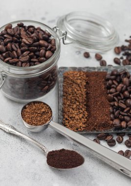 Dondurulmuş hazır kahve taneleri, öğütülmüş kahve ve çelik tabakta fasulyeler, cam kavanoz ve beyaz arka planda çelik kaşıklar..
