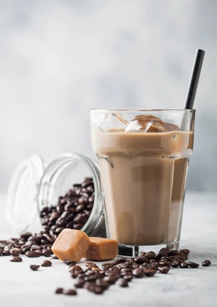 Iced cold coffee with milk in glass with straw and jar of coffee beans with salted caramel and silver scoop on light background.