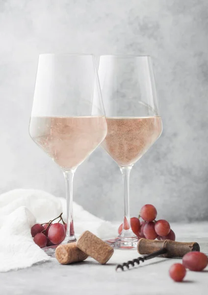 Glasses of rose pink summer wine with corks, grapes and corkscrew on light table background.