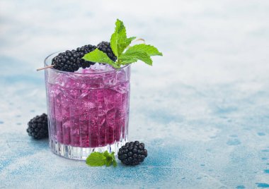 Yaz kokteylini kristal bardakta böğürtlenli, buz küplü ve mavi arka planda nane şekerli tazeliyorum. Soda ve alkol karışımı. Metin için boşluk