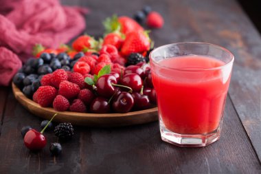 Taze organik yaz meyveleri yuvarlak ahşap tepside ve koyu ahşap masa arkasında bir bardak meyve suyu ile karıştırılır. Ahududu, çilek, yabanmersini, böğürtlen ve kiraz. Üst görünüm