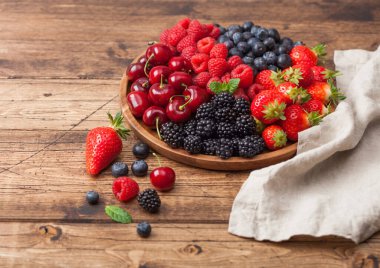 Taze organik yaz meyveleri ahşap masa arka planında yuvarlak ahşap tepside karıştırılır. Ahududu, çilek, yabanmersini, böğürtlen ve kirazlı keten mutfak havlusu. Üst görünüm