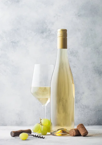 Glass and bottle of summer white wine with grapes, corks and corkscrew on light stone background.