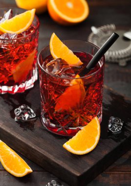 Negroni cocktail in crystal glasses with orange slice and fresh raw oranges on chopping board with strainer on wooden background. Top view