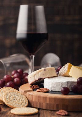 Tahtasında çeşitli peynirler ve ahşap masa zemininde üzümler olan bir bardak kırmızı şarap. Mavi Stilton, Kırmızı Leicester ve Brie Cheese ve bıçak.