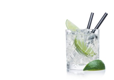 Gimlet cocktail in crystal glass with ice cubes and straw and lime slices on white background with lime on the side. Space for text