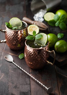 Moscow mule cocktail in a copper mug with lime and mint and wooden squeezer on dark wooden table background and steel shaker and spoon.