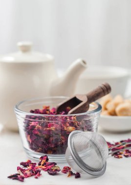 Süzgeç aşılı taze organik gül yaprakları ve beyaz zemin üzerinde beyaz seramik çaydanlık ve baston şekeri..