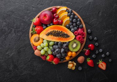 Taze çiğ organik yaz meyveleri ve siyah mutfak arka planında yuvarlak ahşap tabakta egzotik meyveler. Papaya, üzüm, nektarin, portakal, ahududu, kivi, çilek, liçi, kiraz. Üst görünüm
