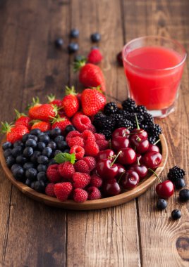 Taze organik yaz meyveleri yuvarlak ahşap tepside ve açık tahta masa arkasında bir bardak meyve suyu ile karıştırılır. Ahududu, çilek, yabanmersini, böğürtlen ve kiraz. Üst görünüm