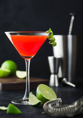 Klasik kristal bardakta kozmopolitan kokteyli, limon kabuğu, süzgeç, jigger ve sallanan taze limonlu siyah masa arkaplanı..