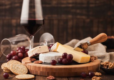Tahtasında çeşitli peynirler ve ahşap masa zemininde üzümler olan bir bardak kırmızı şarap. Mavi Stilton, Kırmızı Leicester ve Brie Cheese ve bıçak.