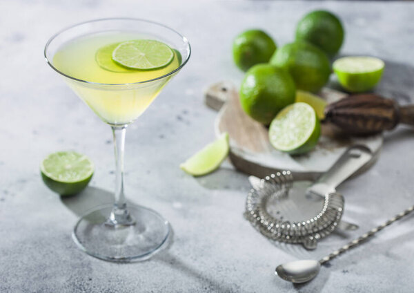 Gimlet Kamikaze cocktail in martini glass with lime slice and ice on light background with fresh limes and strainer with shaker. 