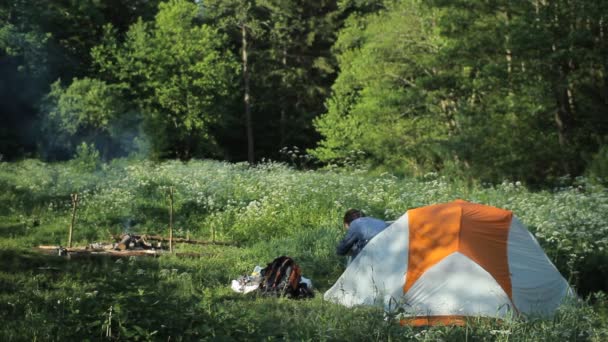 Человек выходит в утренние палатки в лесу. Возле костра курит. Белый человек закрывает палатки и идет к огню — стоковое видео