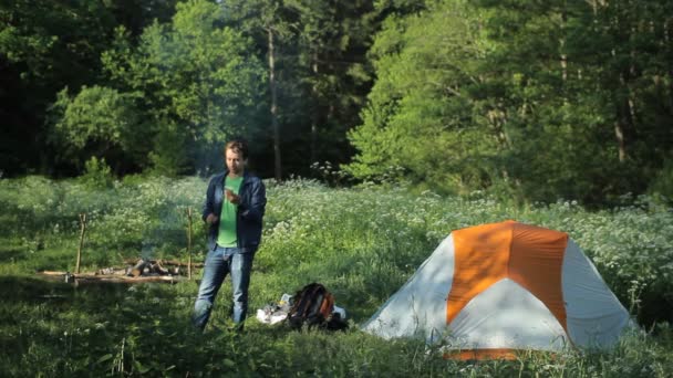 Мужчина разговаривает по телефону у костра и палаток в лесу — стоковое видео