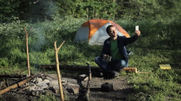 Mannen drikker te ved bålet og tar bilder av skogen og teltet. – stockvideo