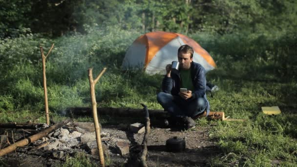 Un hombre bebe té y hace clic en el teléfono cerca de un incendio. En el fondo hay una tienda de campaña y un hermoso bosque — Vídeos de Stock