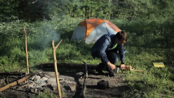 El hombre hace clic en un reloj inteligente y beber té. Cerca del fuego se quema y es tienda en el bosque — Vídeo de stock