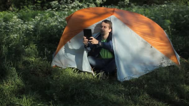 Мужчина фотографирует на планшете рядом с палаткой в лесу — стоковое видео
